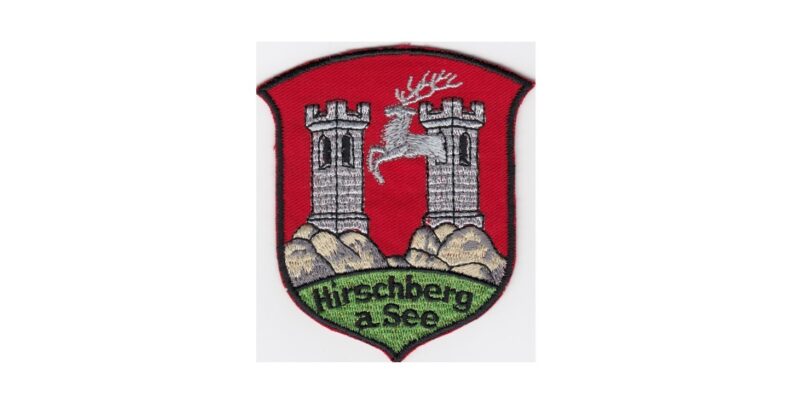 Stadtwappen von Hirschberg (Doksy), Abb. zwei Türme auf roten Untergrund, darunter steht Hirschberg a. See auf grünem Untergrund.